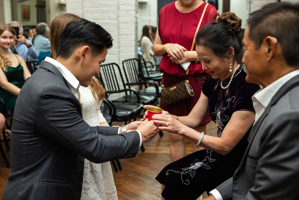 Groom receiving a red gift from smiling older woman during Asian wedding ceremony indoors.