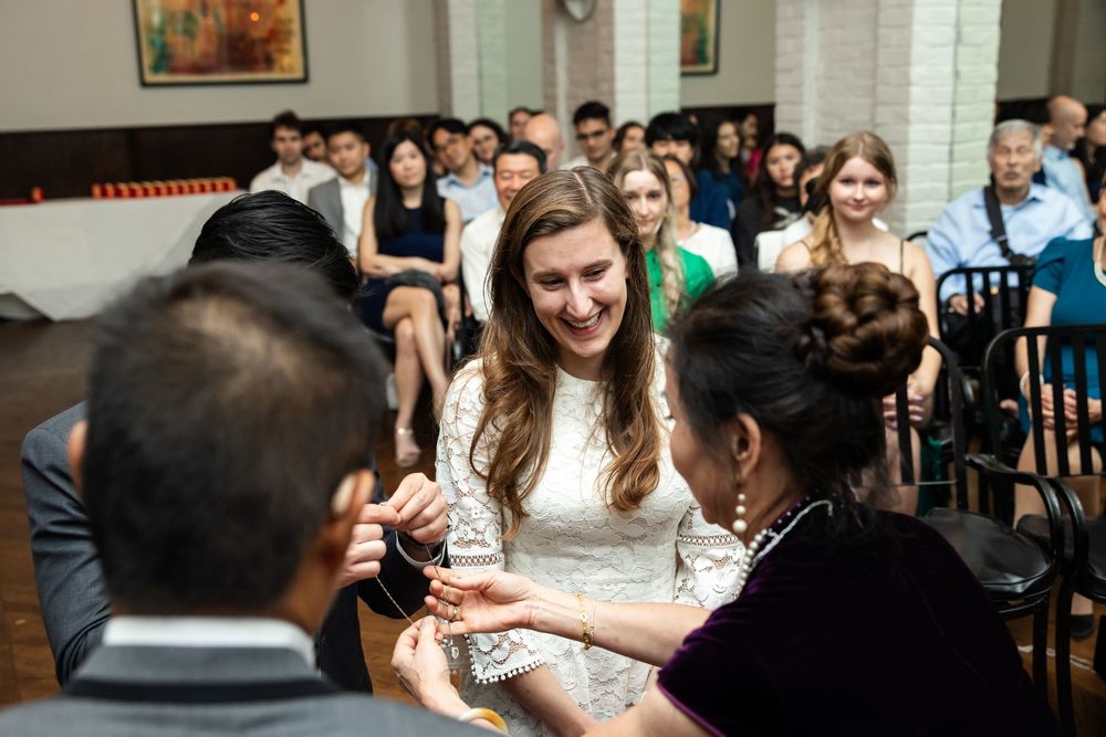 Smiling bride in white lace dress receiving ring during intimate wedding ceremony with seated guests in background.