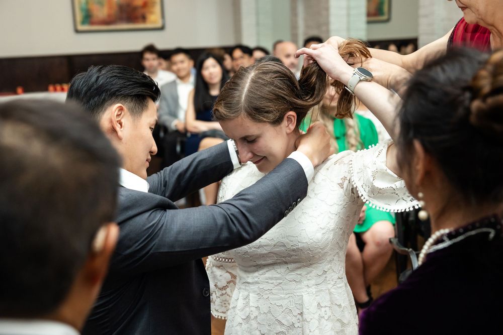 Bride in lace wedding dress receives necklace from groom during emotional wedding ceremony with guests in background.