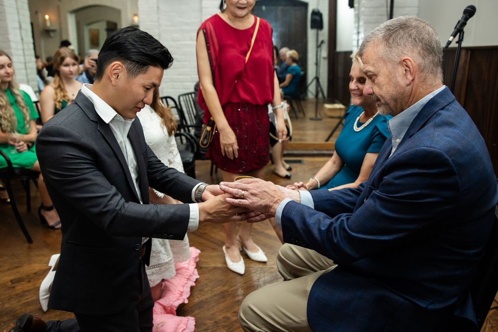 Two men exchange rings during a wedding ceremony indoors, with guests and an officiant standing nearby.