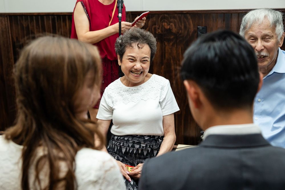 Elderly Asian woman smiling warmly at a couple during a formal indoor gathering with guests in the background.