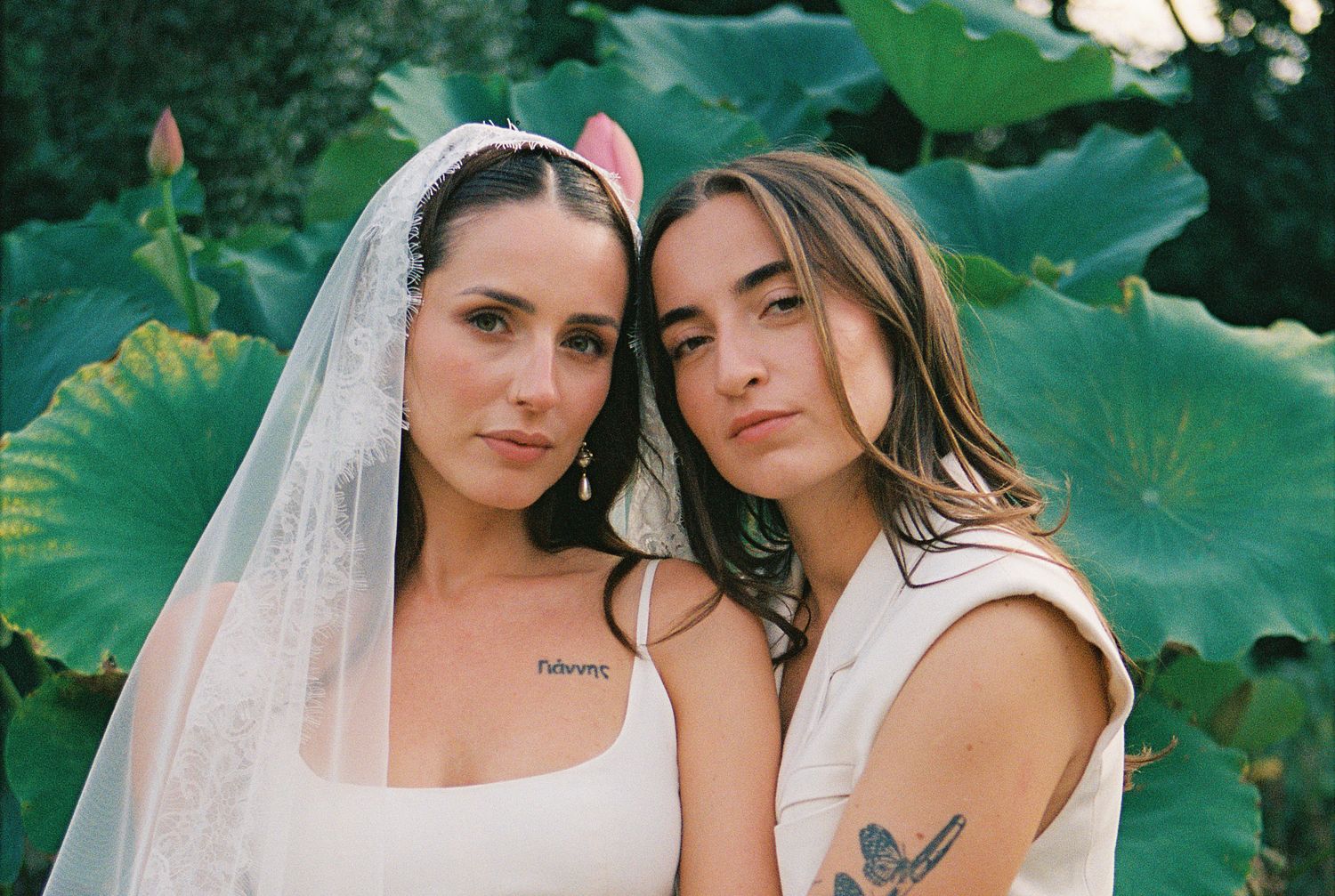 Two brides in white dresses pose together outdoors with lush green tropical foliage in the background.