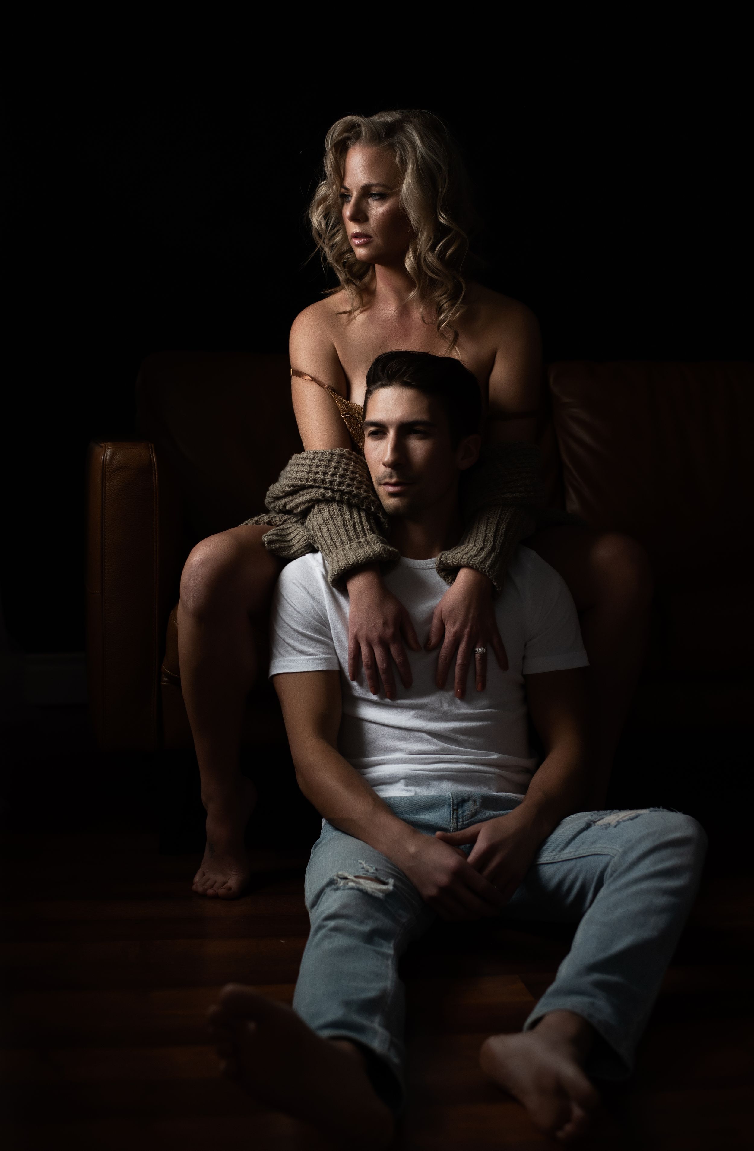 Dramatic moody portrait of a couple in intimate pose on dark leather furniture.