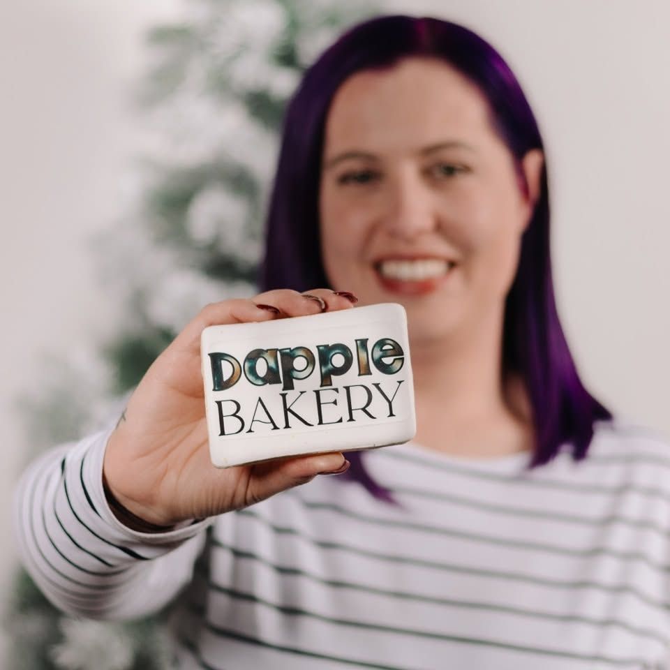 Woman with purple hair smiling and holding a Dapple Bakery gift card in front of a Christmas tree.