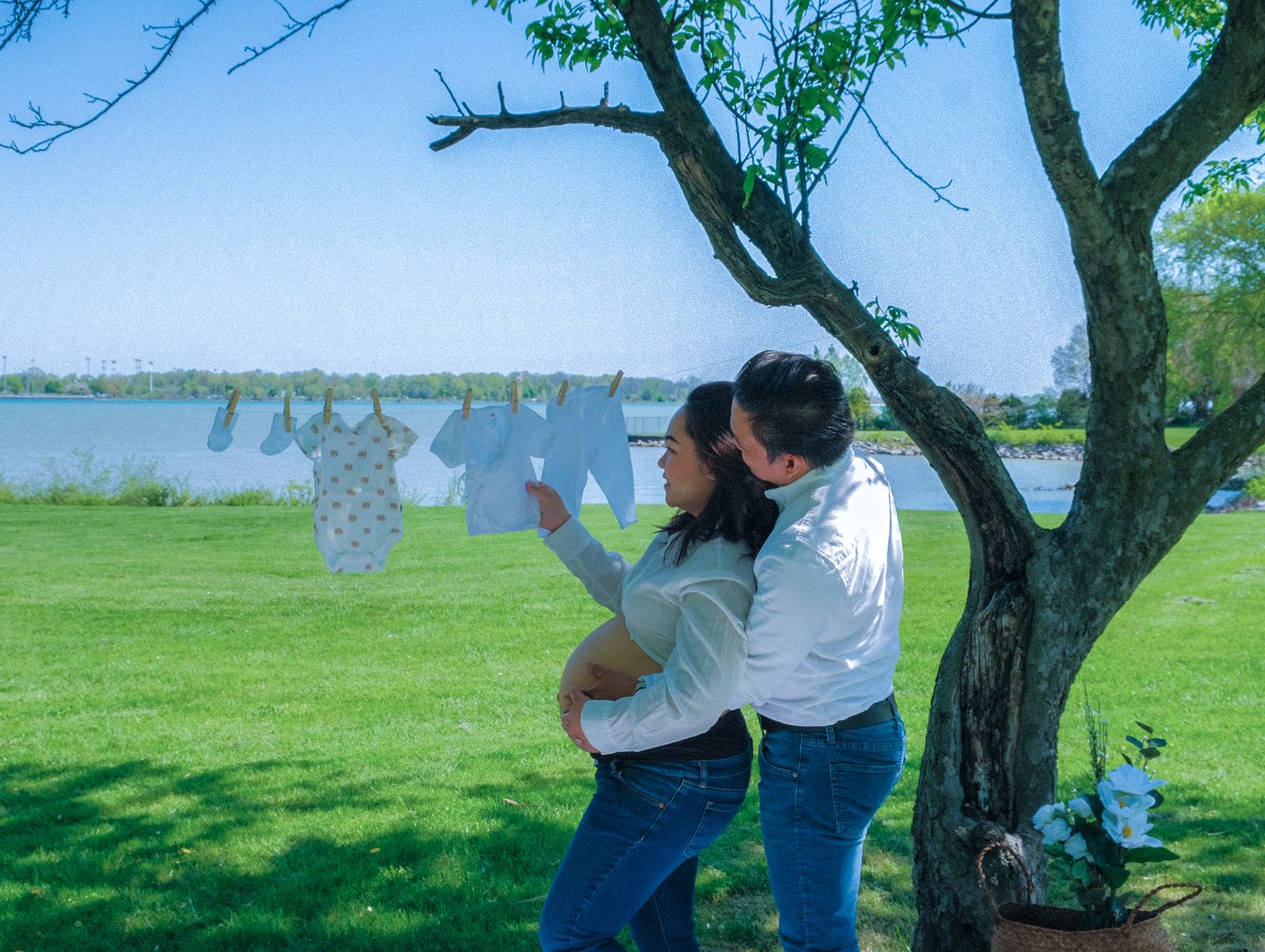 Couple embracing outdoors near a tree with baby clothes on a clothesline, with a serene lake and green lawn in background.