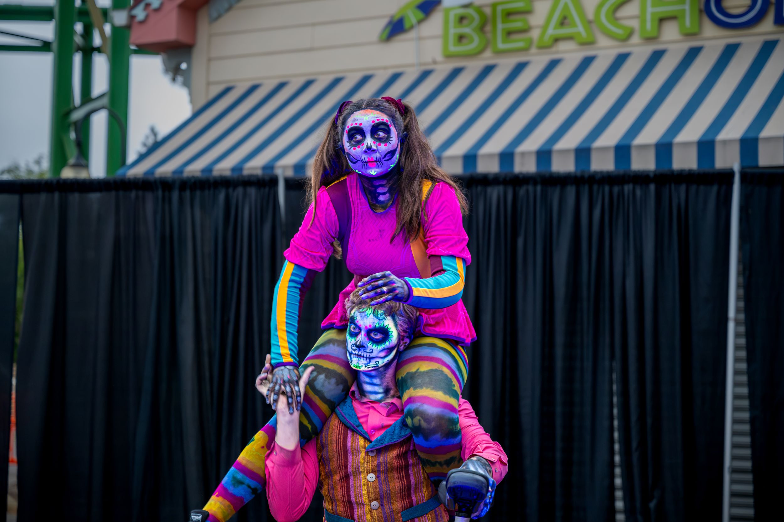 Two performers in colorful Day of the Dead skull makeup and costumes pose at a beach venue Halloween event.