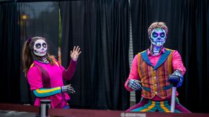 Two performers in colorful Dia de los Muertos skull face paint and costumes pose on stage at a festive event.