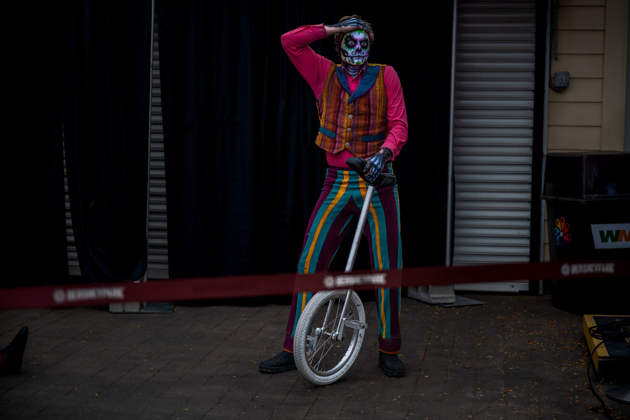 A colorful clown in rainbow striped pants and skull face paint poses with a unicycle outside a circus venue.