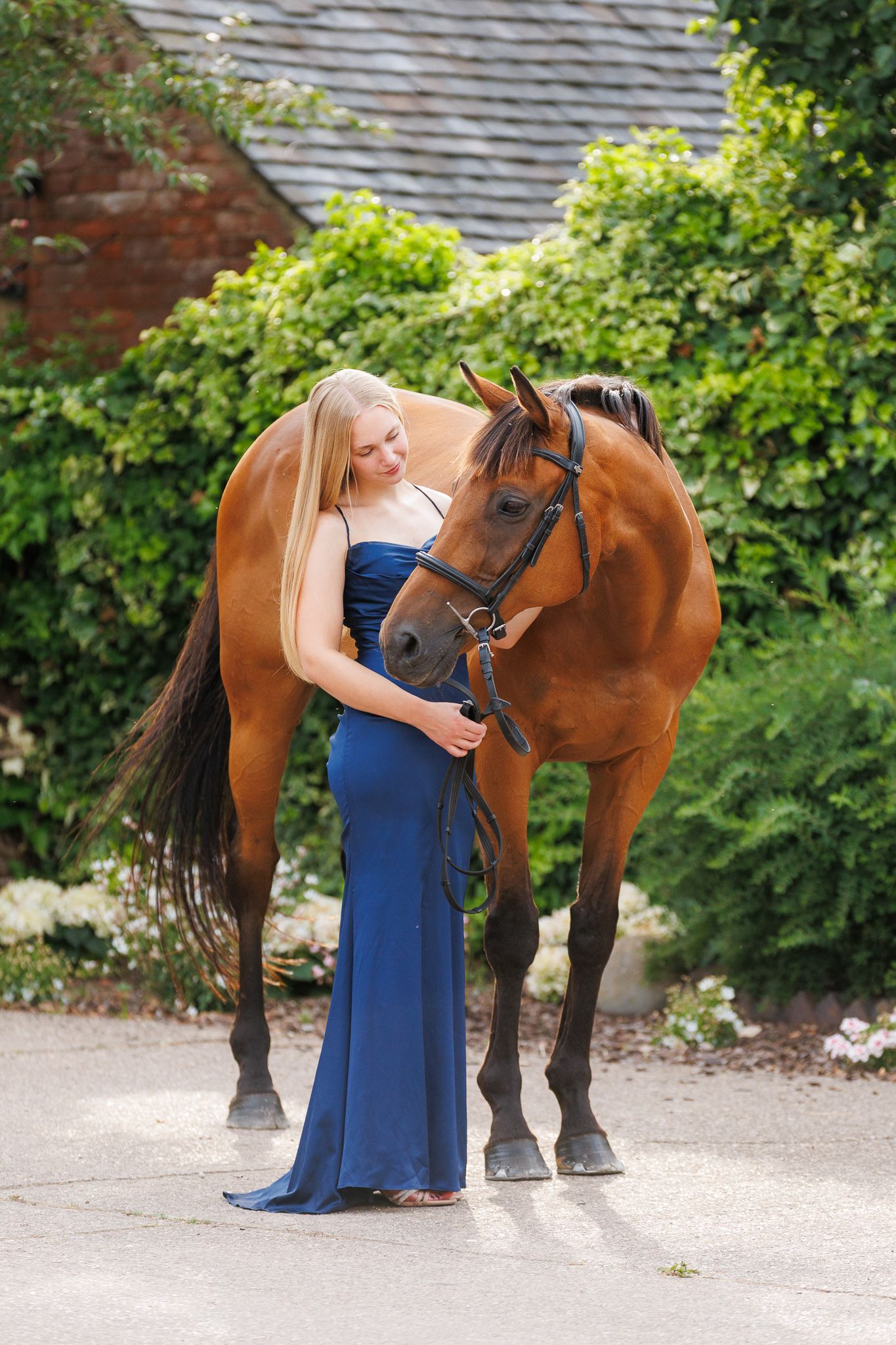 Woman with long blonde hair in a navy blue gown stands beside a bay horse in a bridle, with green hedges and a brick building behind.