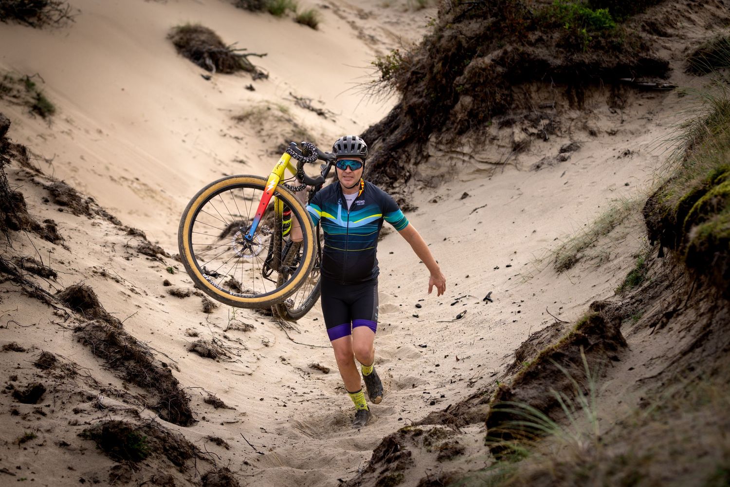 Cyclist in a teal jersey carries a mountain bike on one shoulder while hiking up a steep, sandy dune trail on a sunny day.