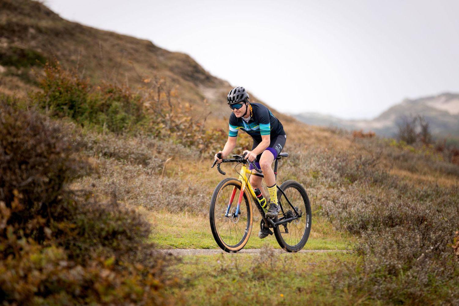 Cyclist in a helmet and sunglasses rides a yellow gravel bike along a grassy trail through rolling dunes and shrubland.