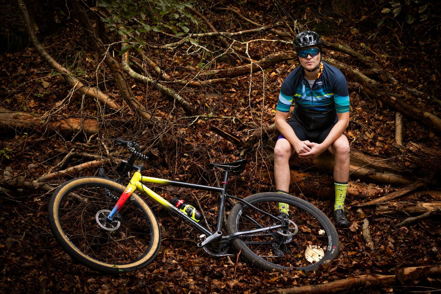 Cyclist in a blue jersey and helmet sits on a fallen log in an autumn forest beside a yellow gravel bike resting on leaves.