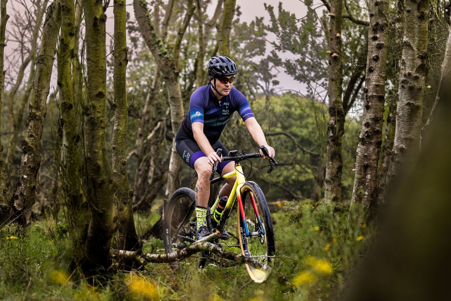Mountain biker in a blue jersey and helmet rides a yellow mountain bike along a forest trail lined with birch trees and wildflowers.