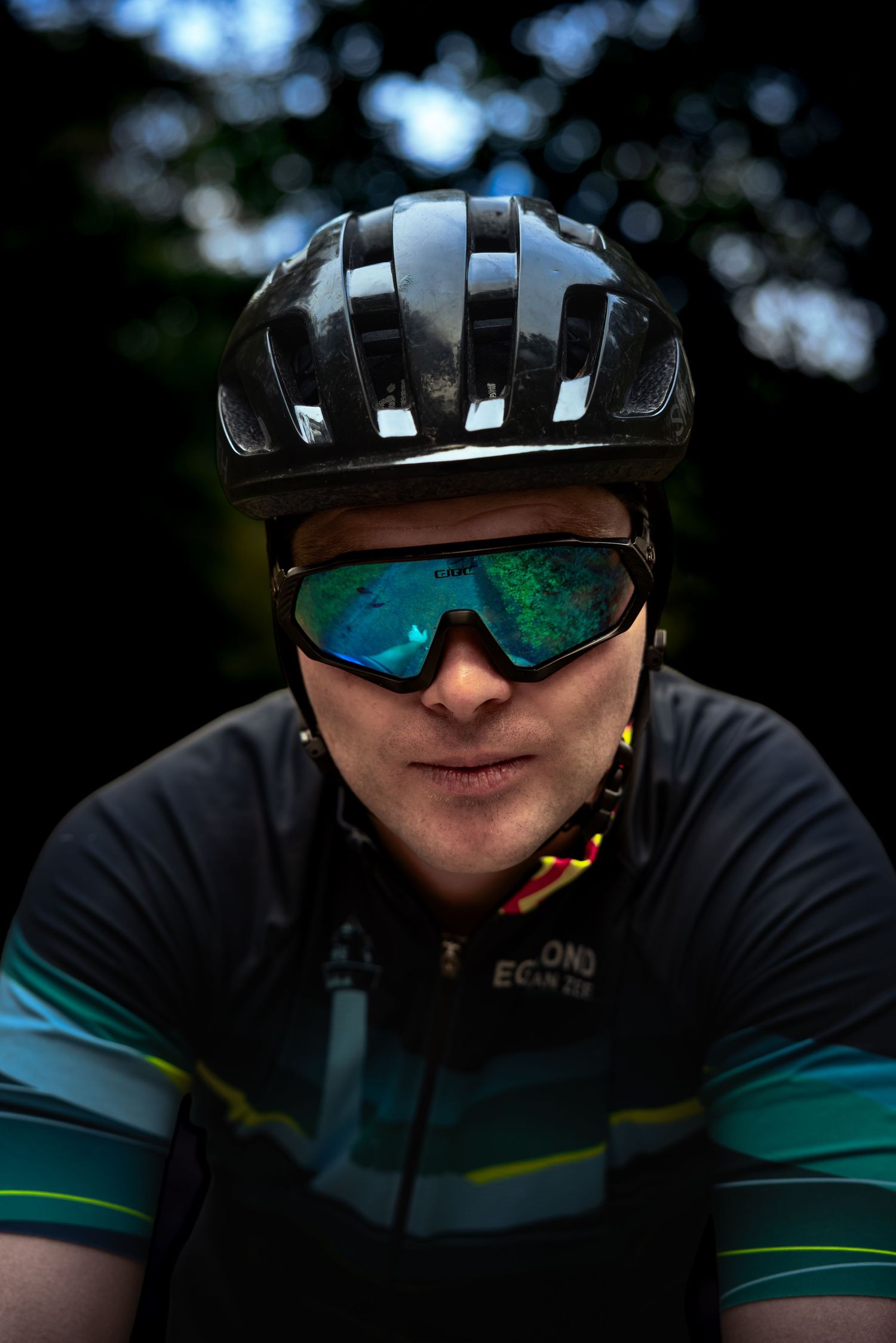 Close-up portrait of a cyclist in a black helmet, green mirrored sport sunglasses and a teal-striped cycling jersey against a dark forest.