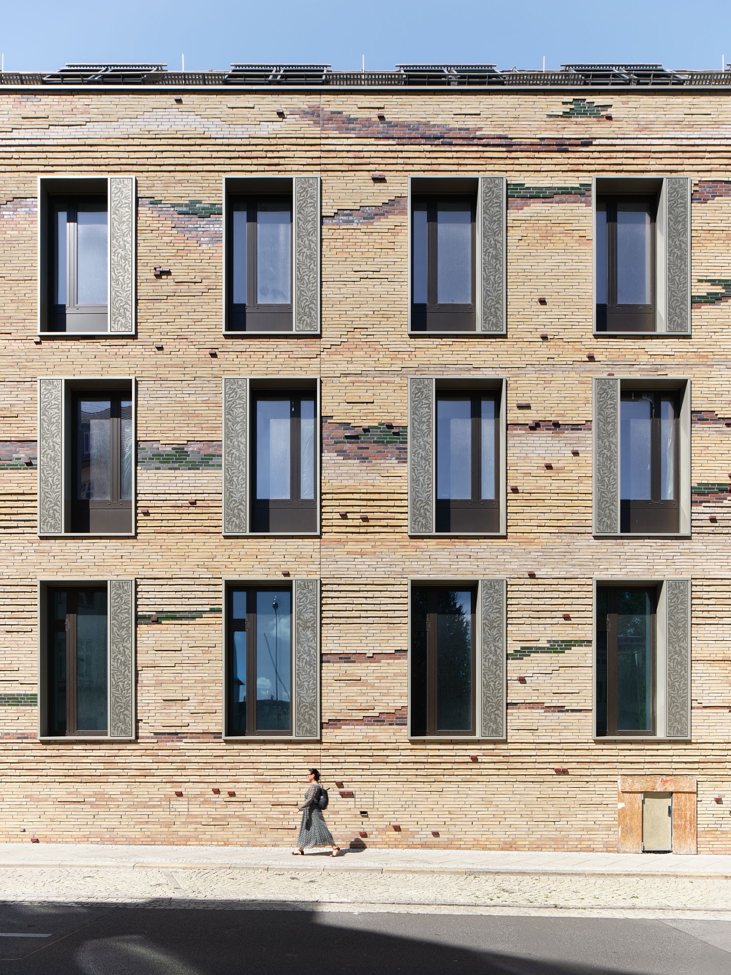 Modern brick apartment facade with multicolored textured bricks and deep-set windows with gray shutters as a pedestrian walks past.