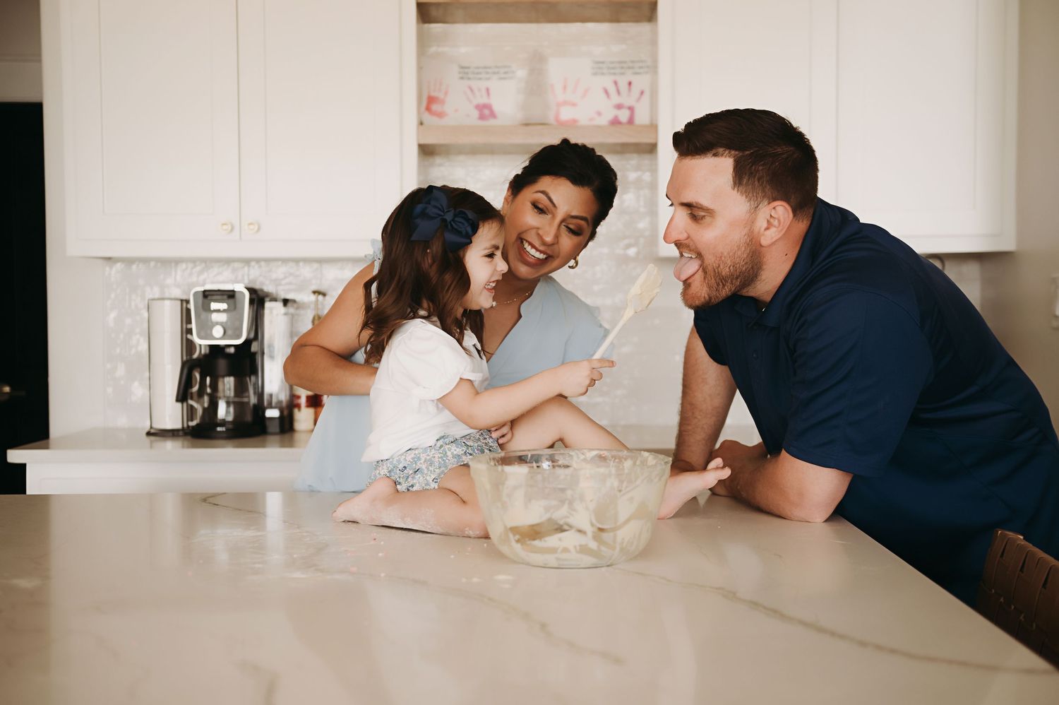 Smiling family of three baking together in a bright white kitchen as a young girl sits on the counter and stirs batter in a glass bowl.