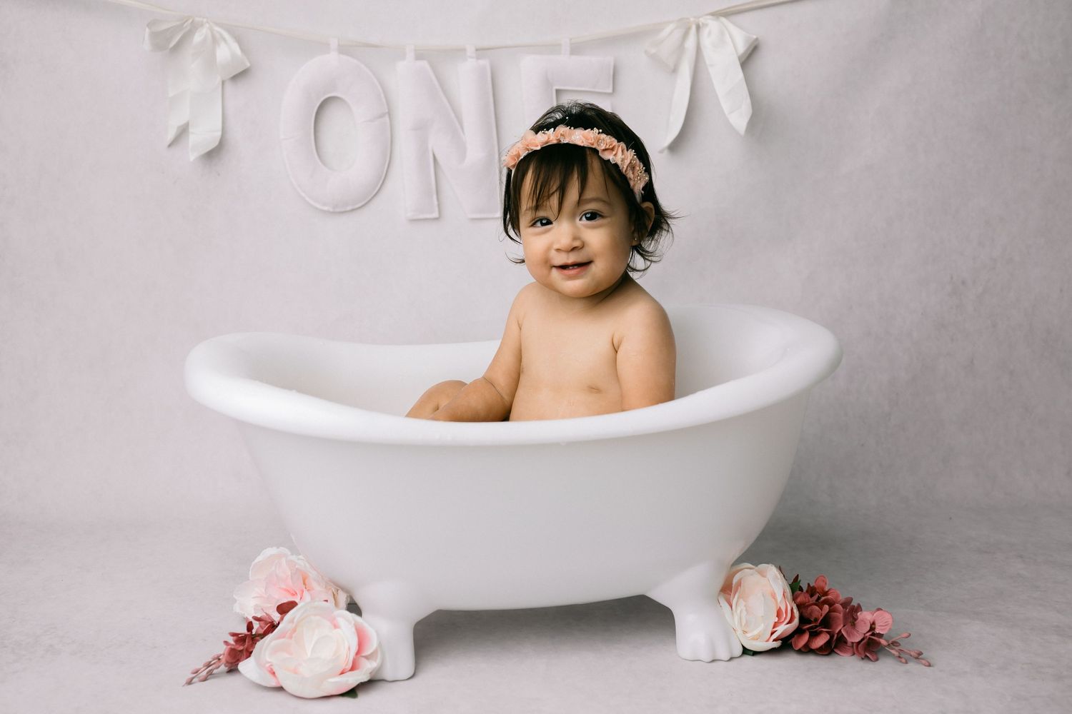 Baby girl in white bathtub smiling, wearing floral headband, ONE banner behind her, pink roses, first birthday photo.