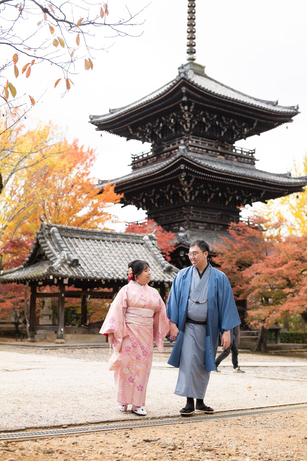 Couple in traditional Japanese kimono walk hand in hand past a pagoda and temple pavilion surrounded by colorful autumn foliage.