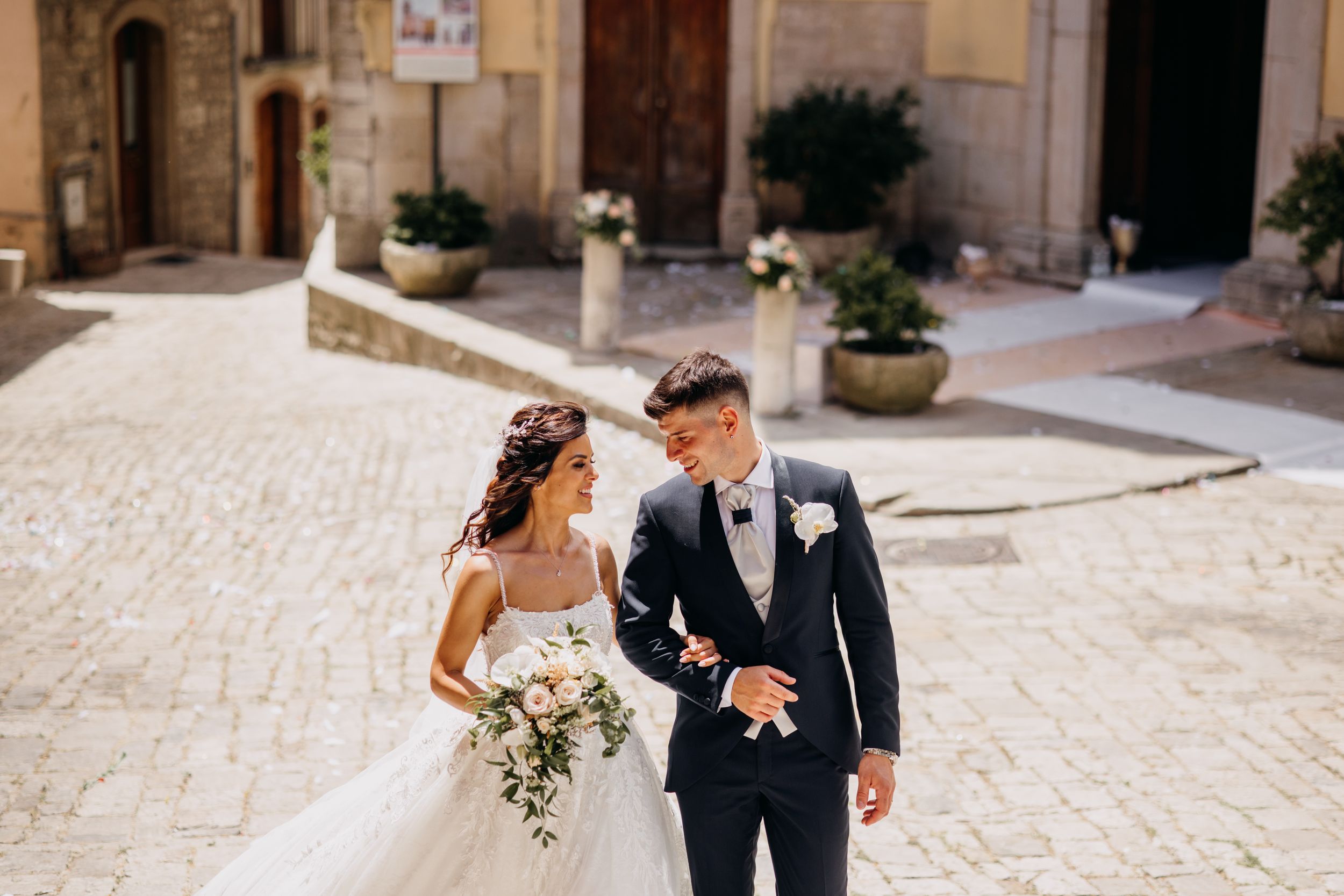Bride and groom walk hand in hand across cobblestone plaza outside historic church on wedding day.
