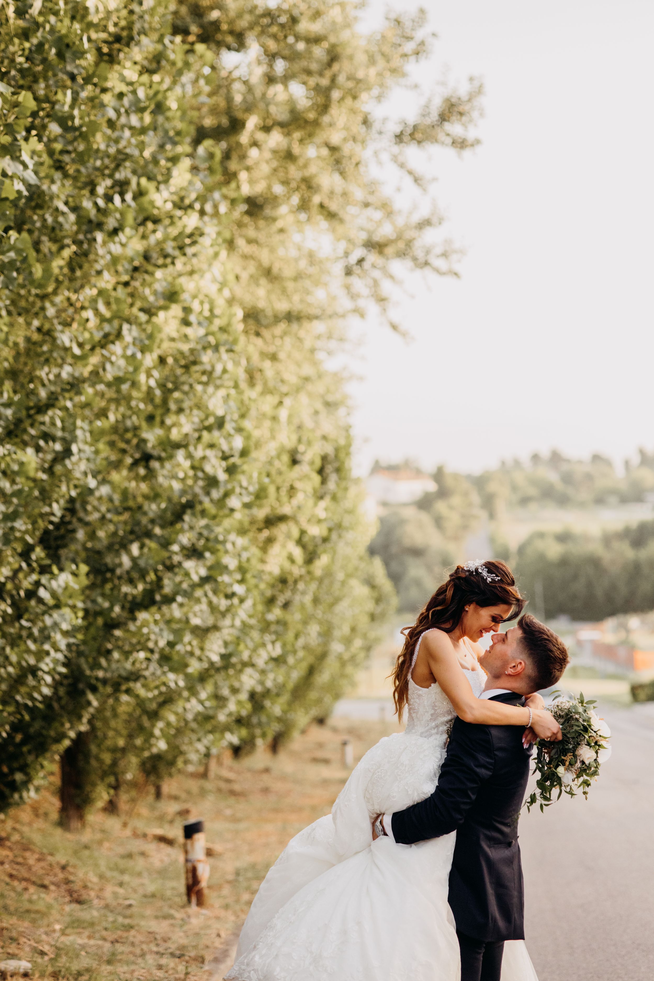 Groom lifting bride in a white gown along a tree-lined road during outdoor wedding photos.