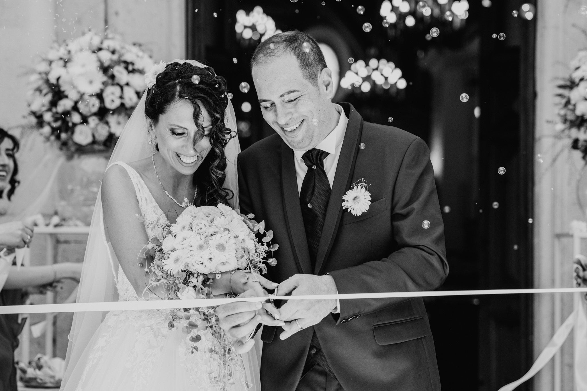 Black and white photo of a laughing bride and groom cutting a ribbon outside a church with bubbles.