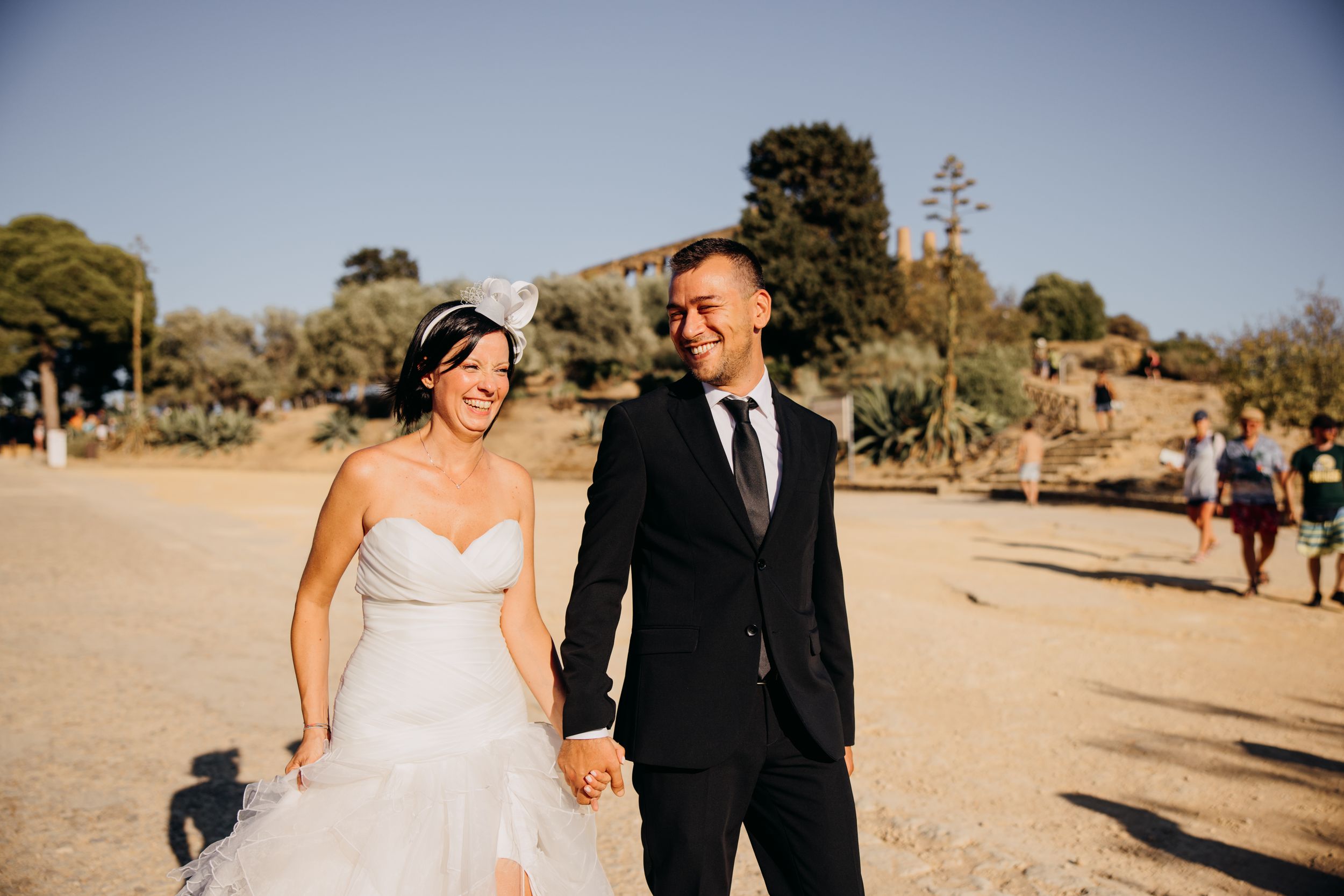 Smiling bride and groom holding hands walking in sunlight at an outdoor wedding venue.