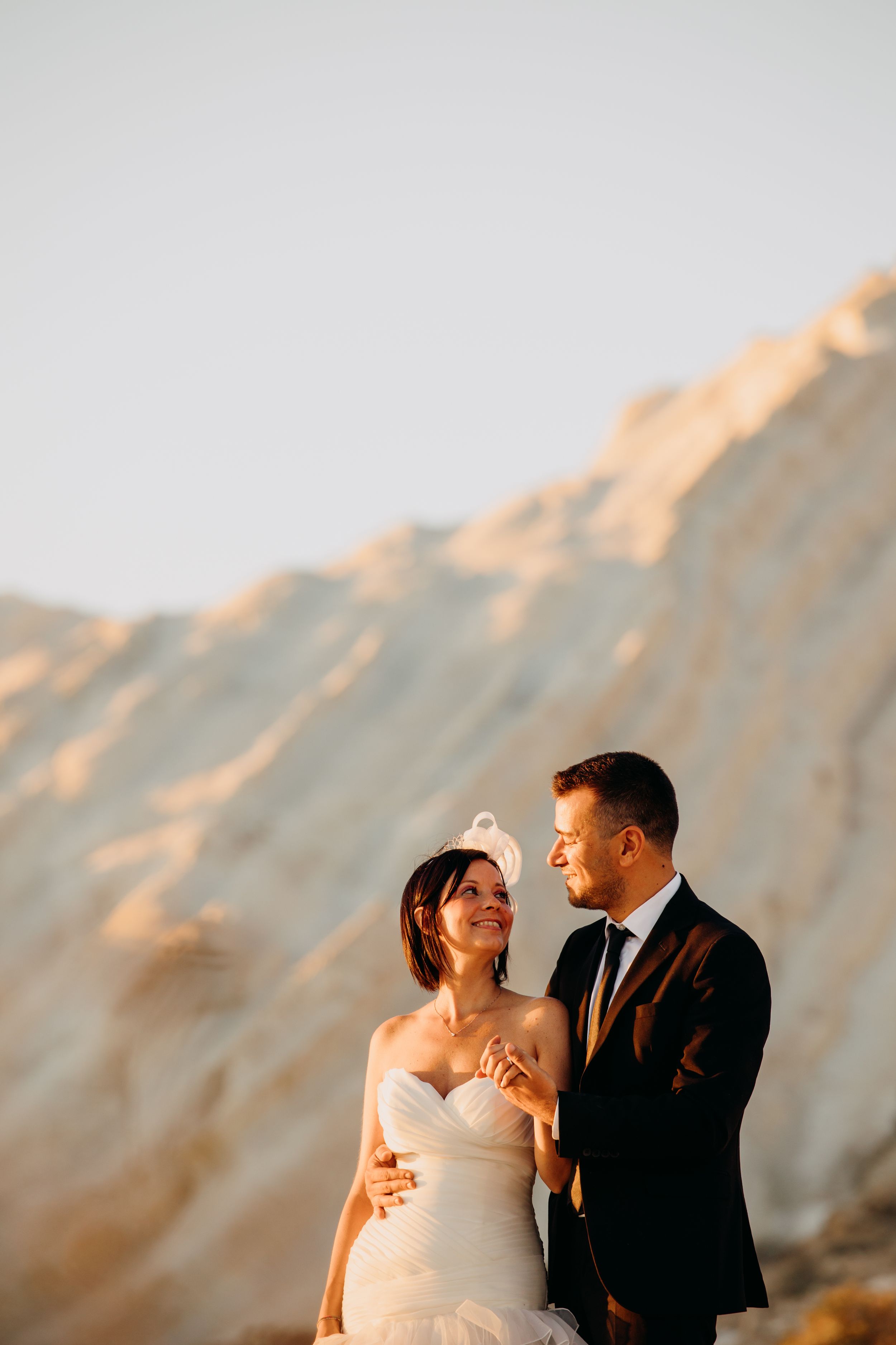Bride and groom holding hands and smiling before golden sandy cliffs at sunset.