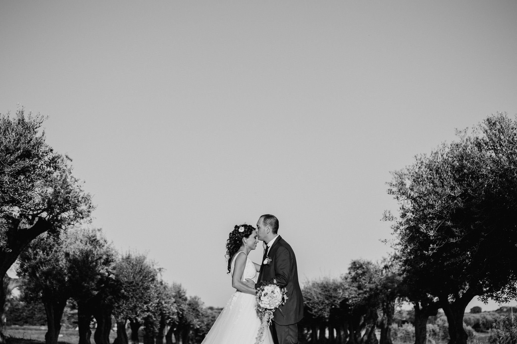 Black and white wedding photo of bride and groom kissing between trees in an open field.
