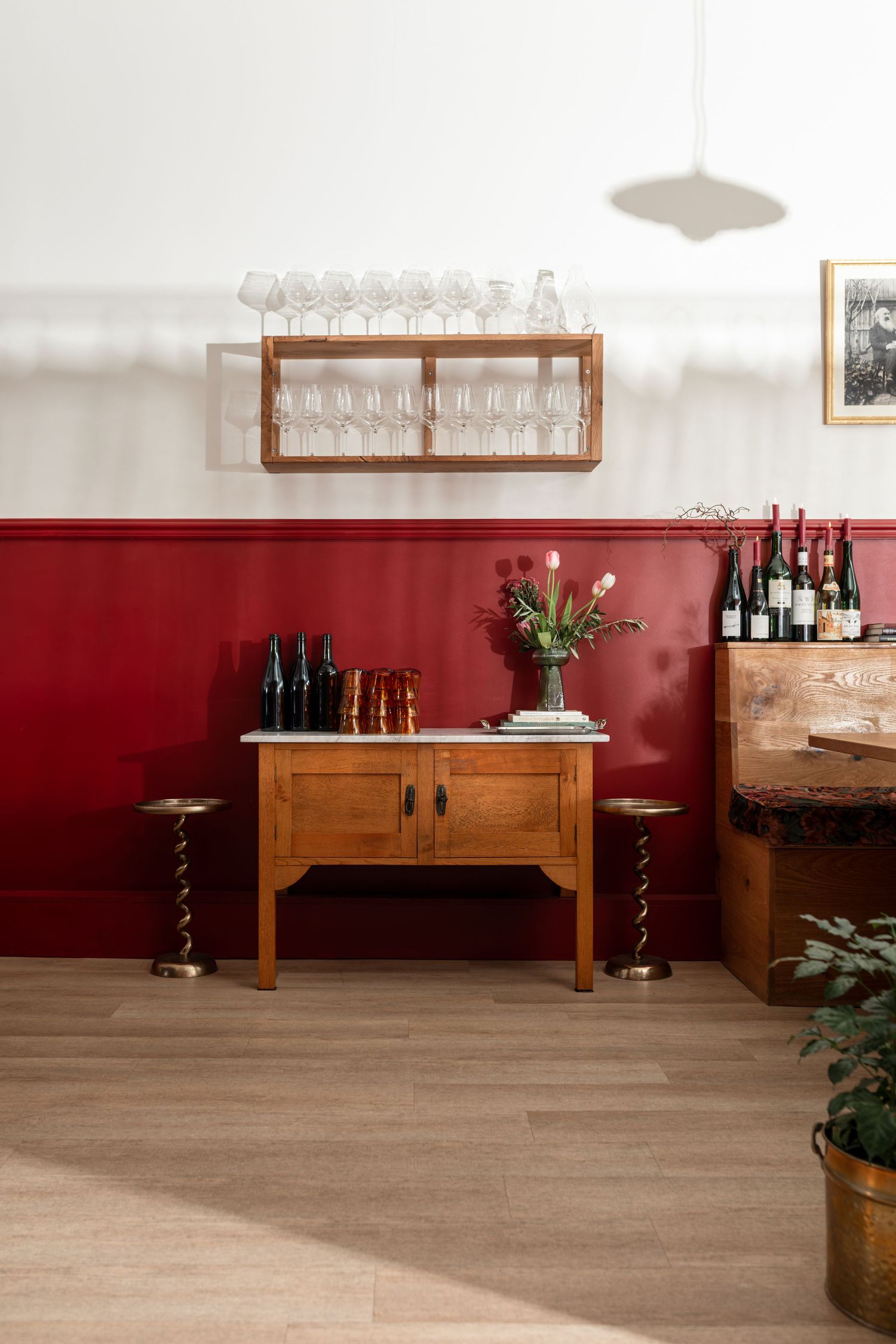 Wooden sideboard with wine bottles and tulips against a red wainscot wall, with a glassware shelf above and twisted wooden stools.
