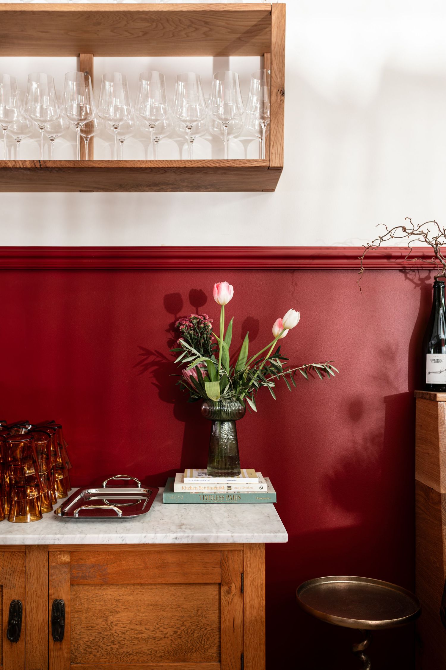 Pink tulips in a dark green vase on a marble-topped wooden cabinet against a burgundy wall, with wine glasses on a shelf above.