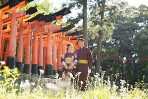 A couple in traditional Japanese kimonos stands among wildflowers in front of the orange torii gates of a shrine surrounded by trees.