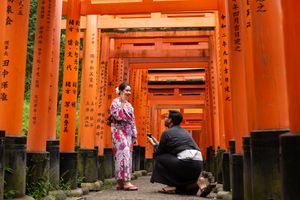 A woman in a floral kimono stands among vermilion torii gates while a seated man takes a photo at Fushimi Inari Shrine in Kyoto.