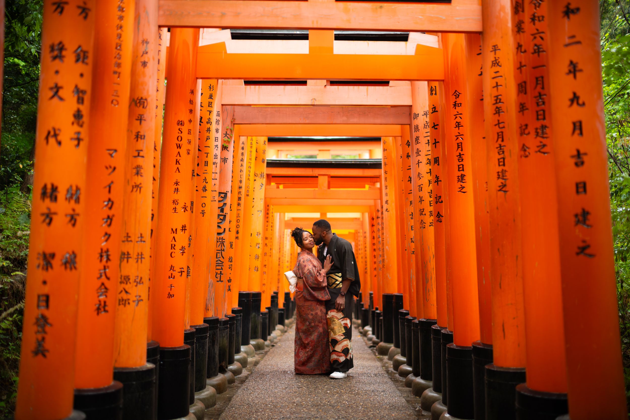 Couple in traditional dress embracing in a corridor of orange torii gates at a Japanese shrine, with black kanji inscriptions.