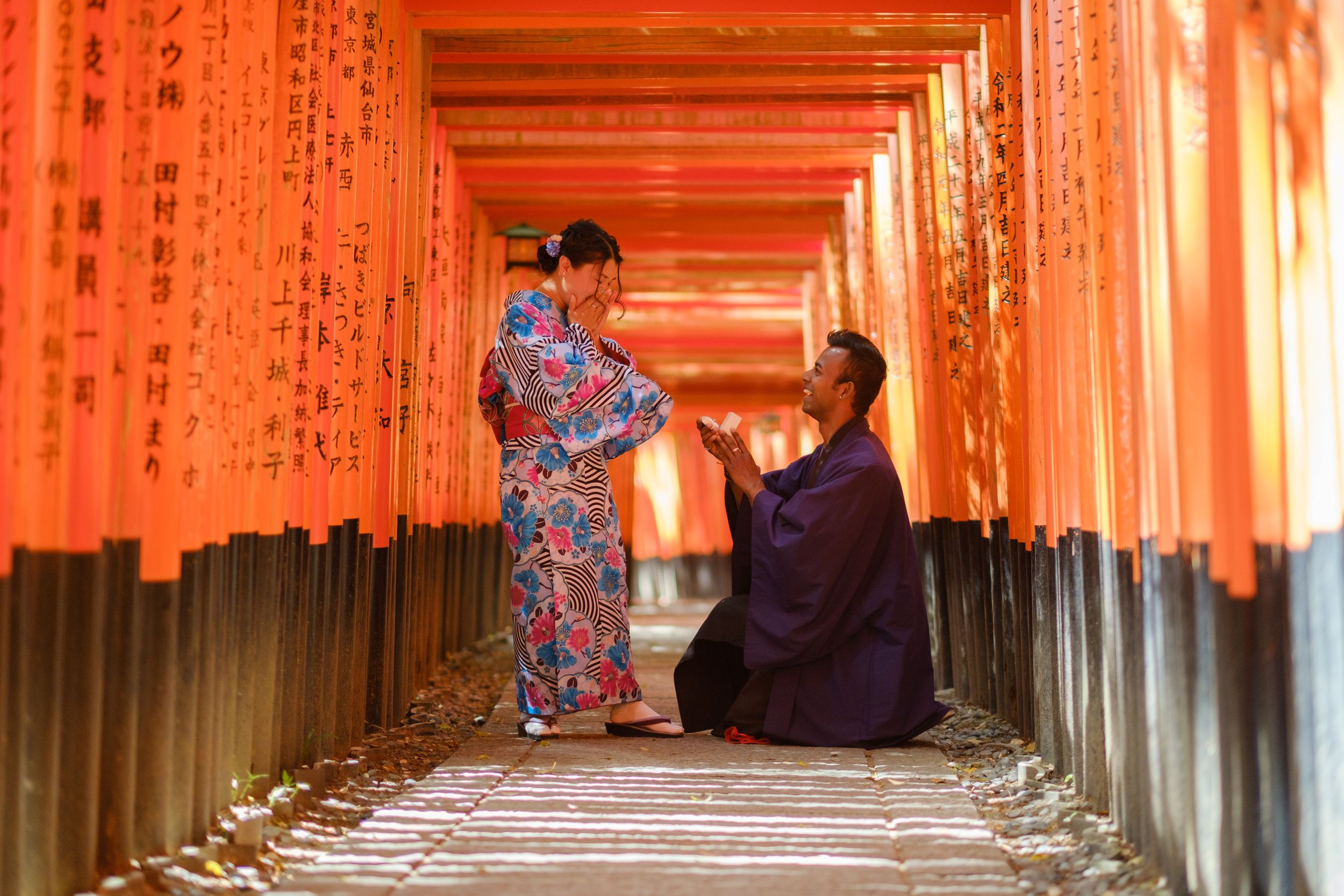 A man in a dark kimono kneels and gestures toward a woman in a floral kimono among the red torii gates at Fushimi Inari shrine in Kyoto.