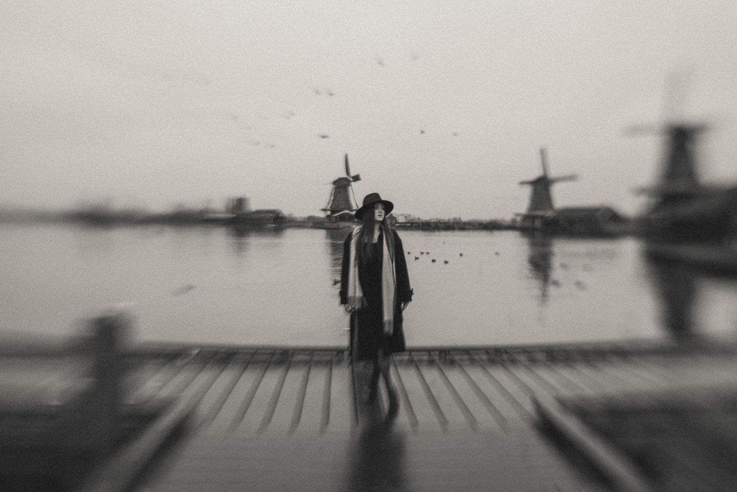 Woman in a wide-brimmed hat standing on a wooden pier by a calm lake with traditional Dutch windmills in the misty background. Zaanse Schans