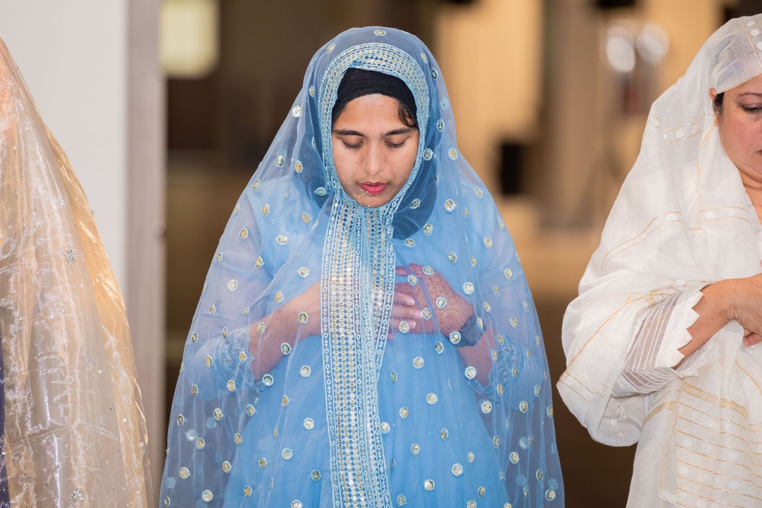 Muslim Woman in a sheer blue embroidered headscarf and gown with silver beadwork prays with hands clasped beside a person in white.