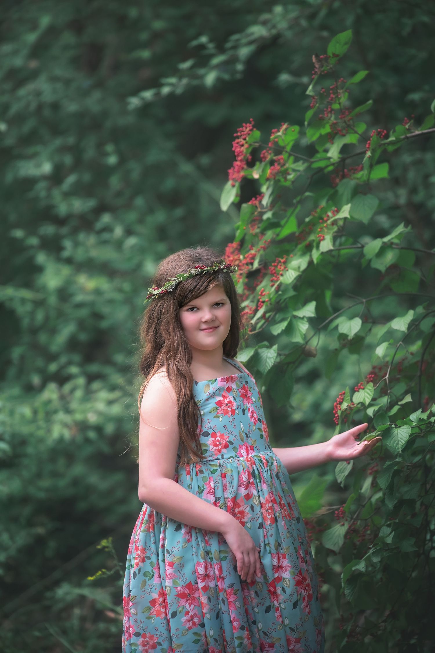 Girl in a teal floral dress and flower crown stands in a lush green forest, gently touching leaves of a bush with red blossoms.