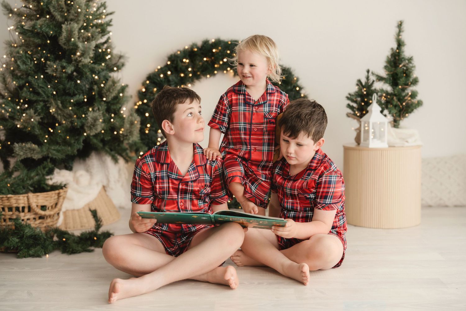 Three children in matching red plaid pajamas read a green storybook together beside a lit Christmas tree and holiday wreath.