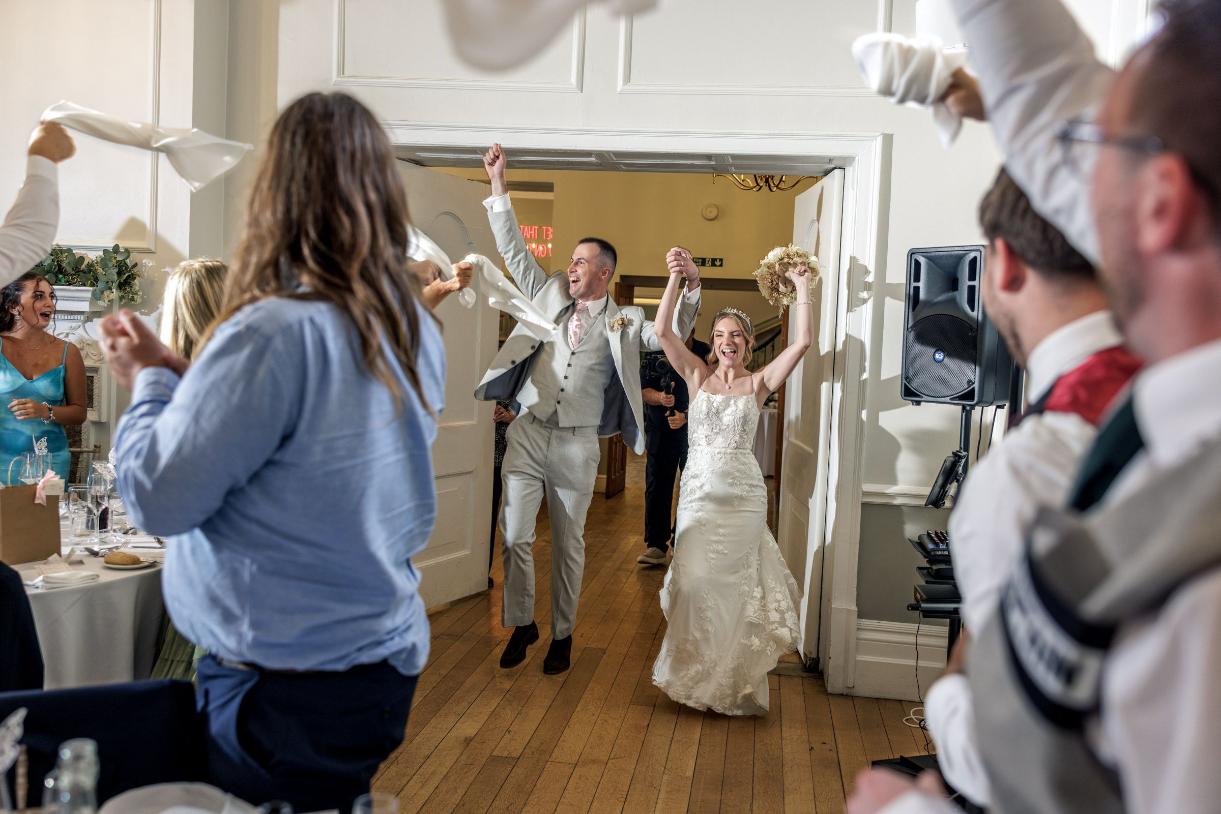 Bride in a white gown and groom in a gray suit dance joyfully into the reception as cheering wedding guests raise their arms.