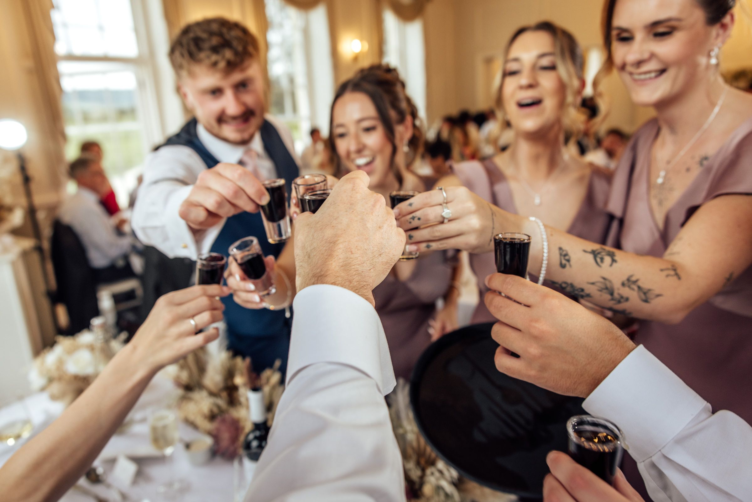 Smiling wedding guests clink small glasses together in a joyful toast at a festive reception inside a bright, elegant venue.