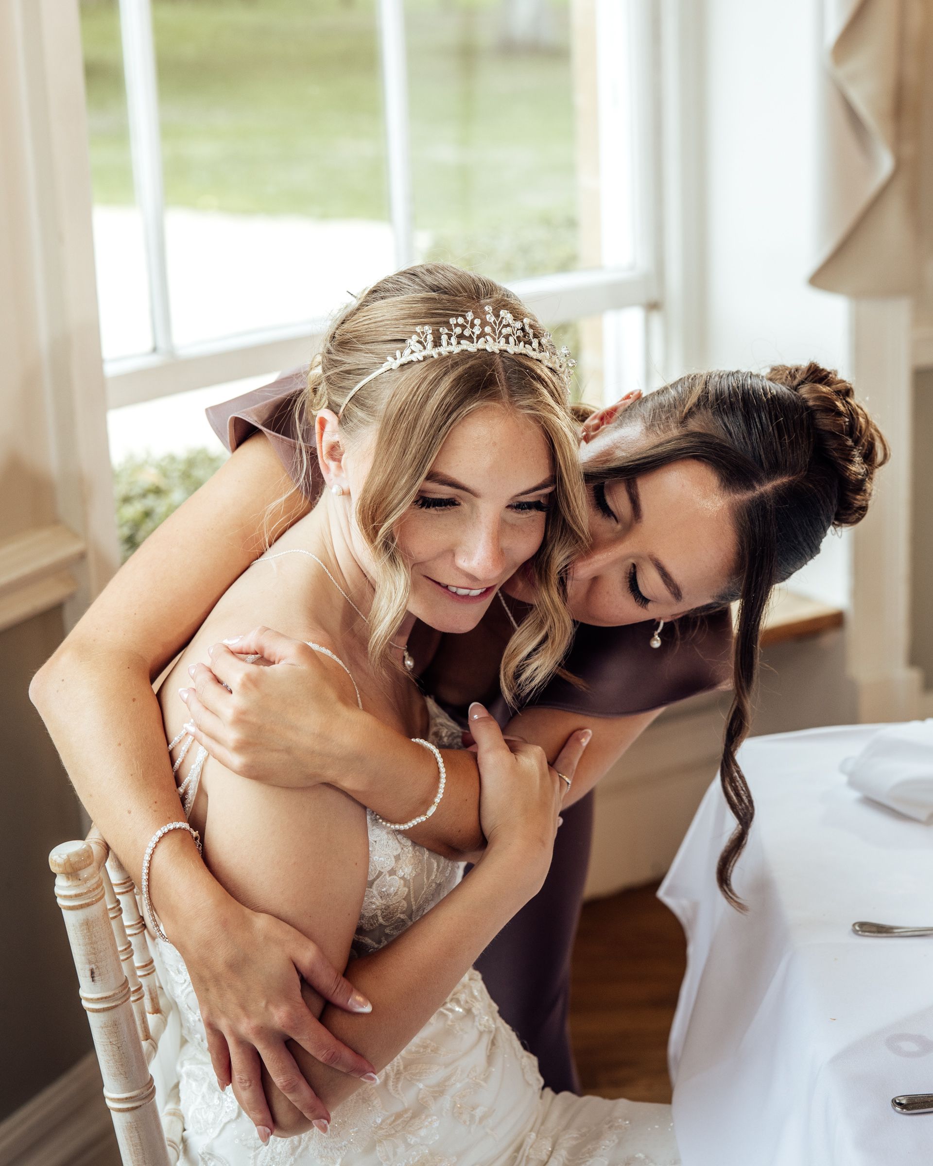 A bride in a lace wedding gown and tiara smiles warmly as a bridesmaid in a plum dress hugs her from behind at a reception table.