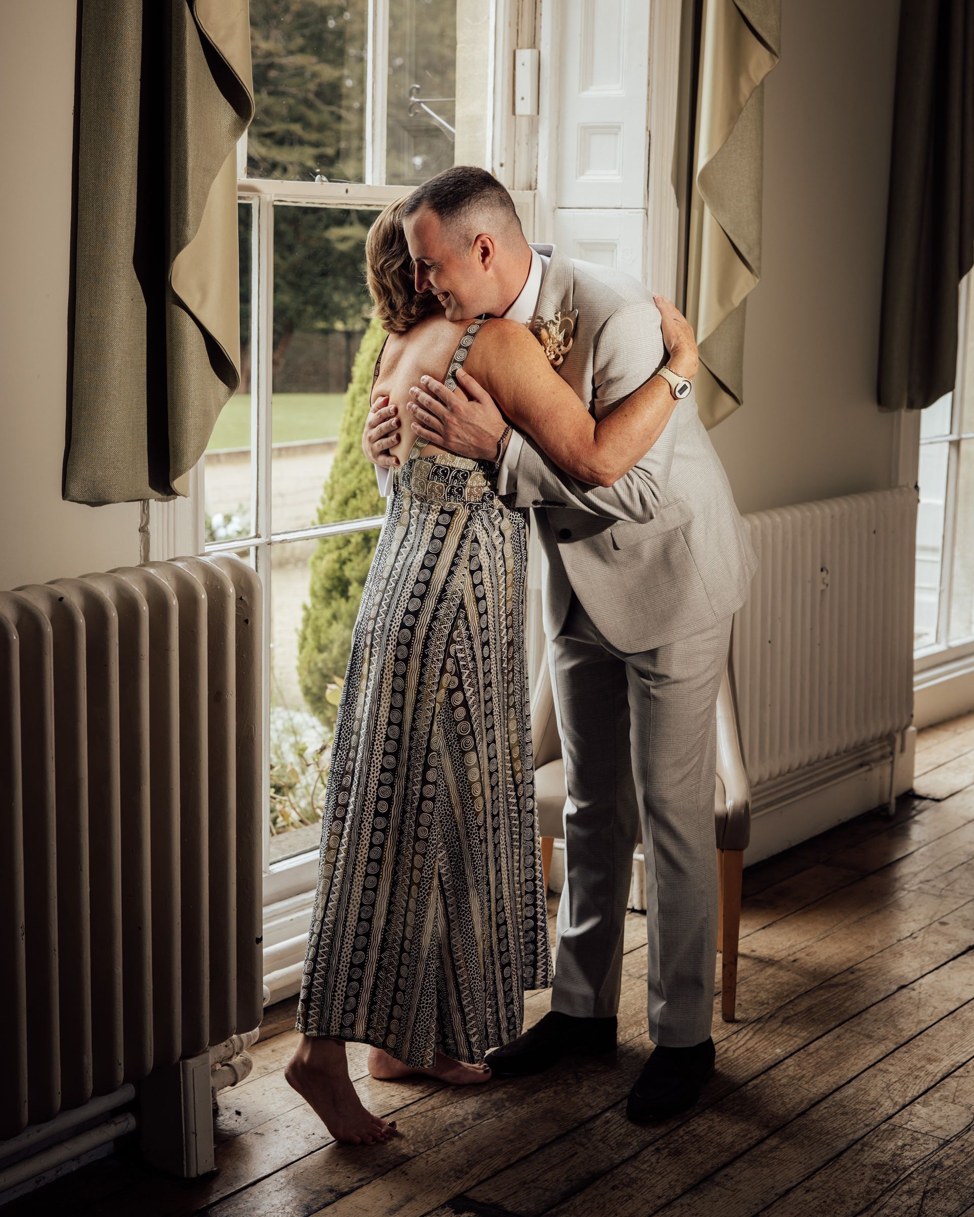 A groom in a gray suit tenderly embraces a barefoot woman in a patterned maxi dress beside a bright window in a rustic room.