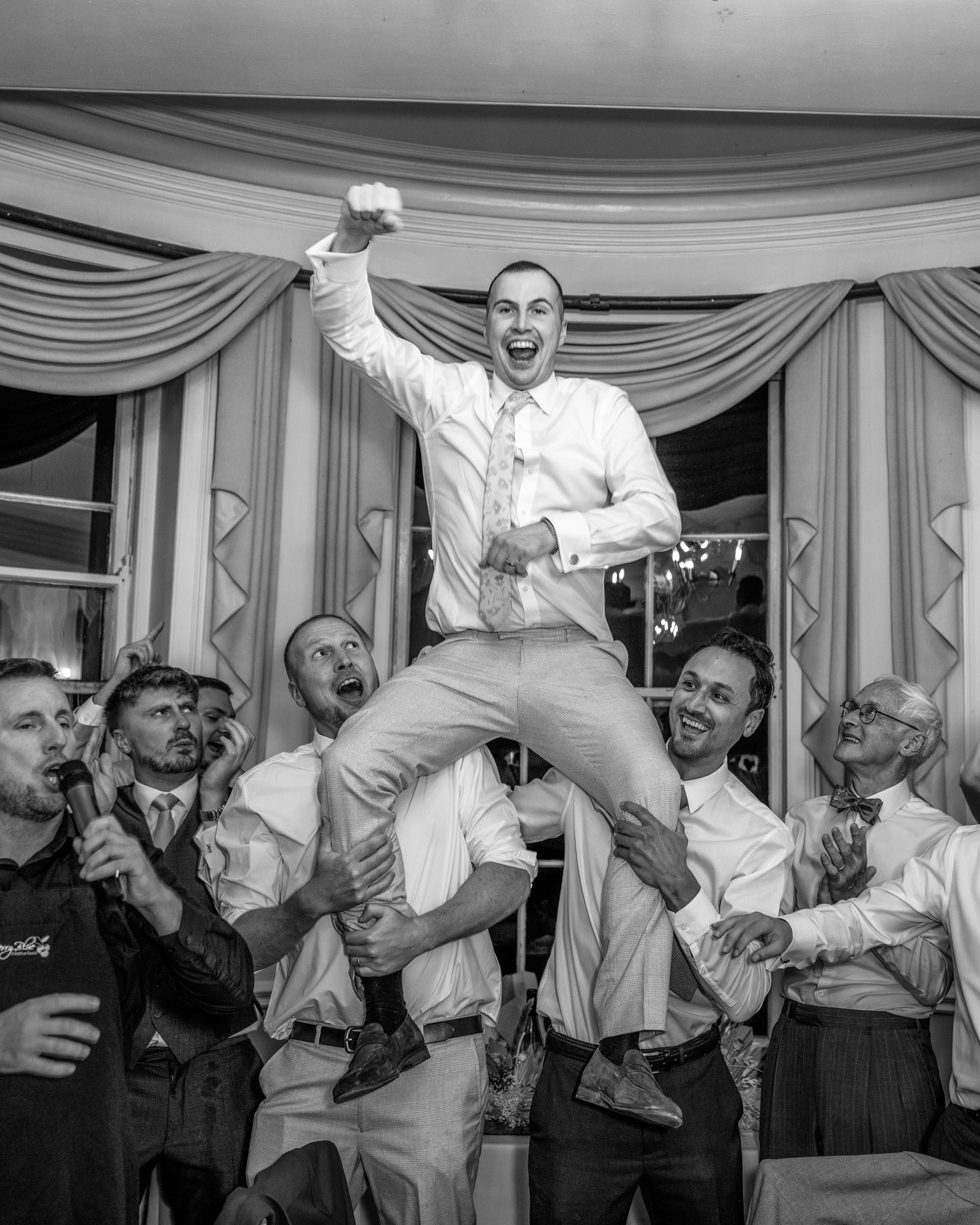 Black and white photo of wedding guests lifting a cheering man in the air on their shoulders during a lively reception celebration.