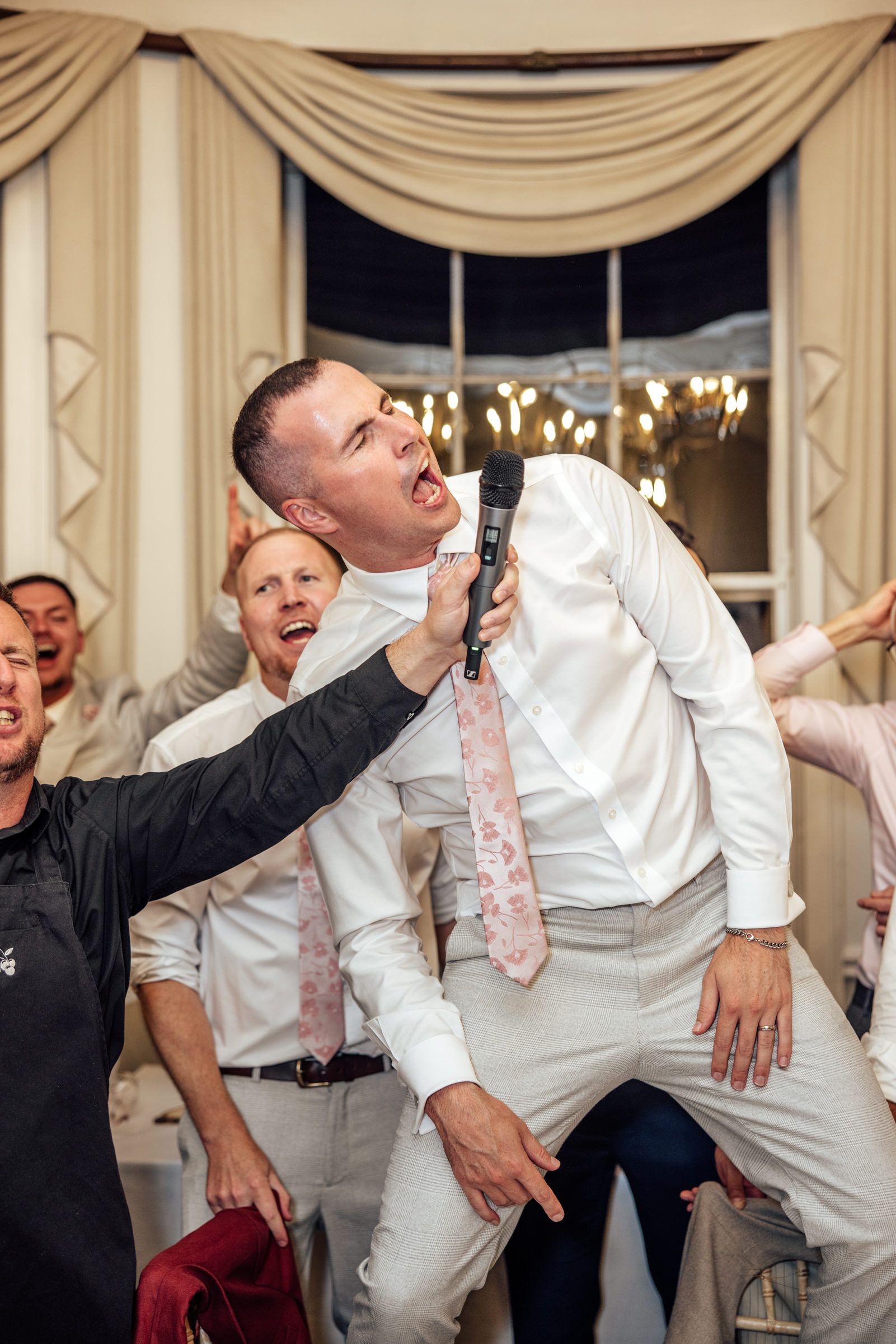 Groomsman in a white shirt and pink floral tie leans back singing into a microphone as friends cheer at a wedding reception.