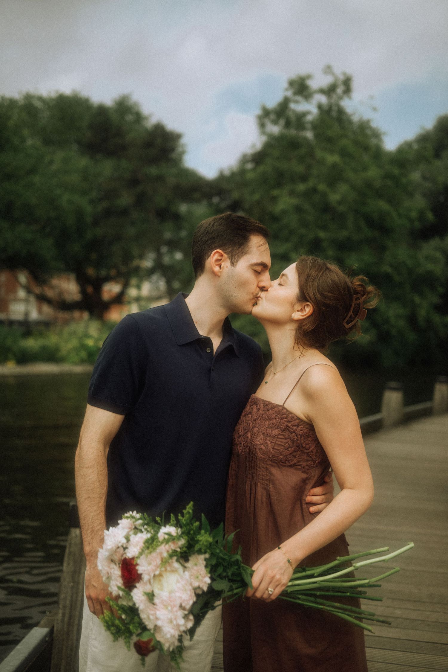 Couple kissing on a wooden dock by the water, the woman holding a bouquet of white and red flowers, Amsterdam.