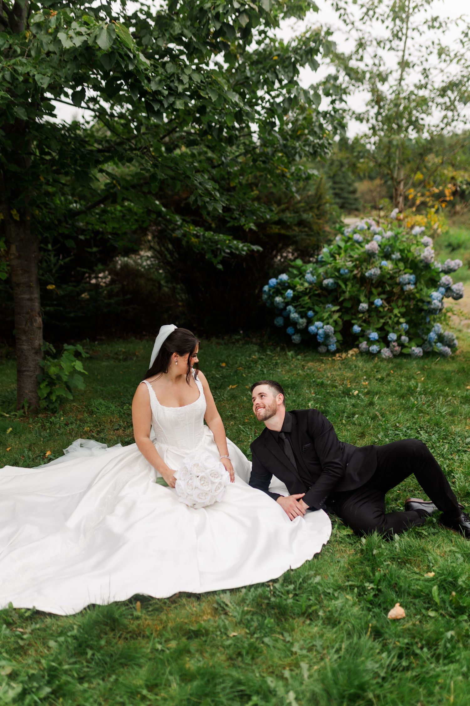 Bride in a white gown sits on green grass beside a groom in a black suit, with blue hydrangeas and trees in the background.