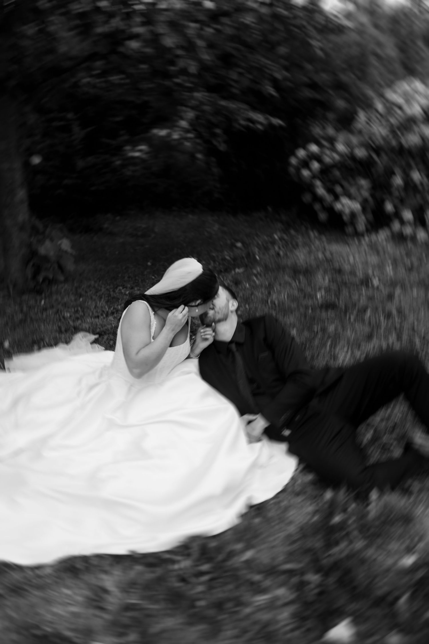 Black and white photo of a bride in a white gown and a groom in a dark suit sharing a romantic kiss while seated on the grass outdoors.