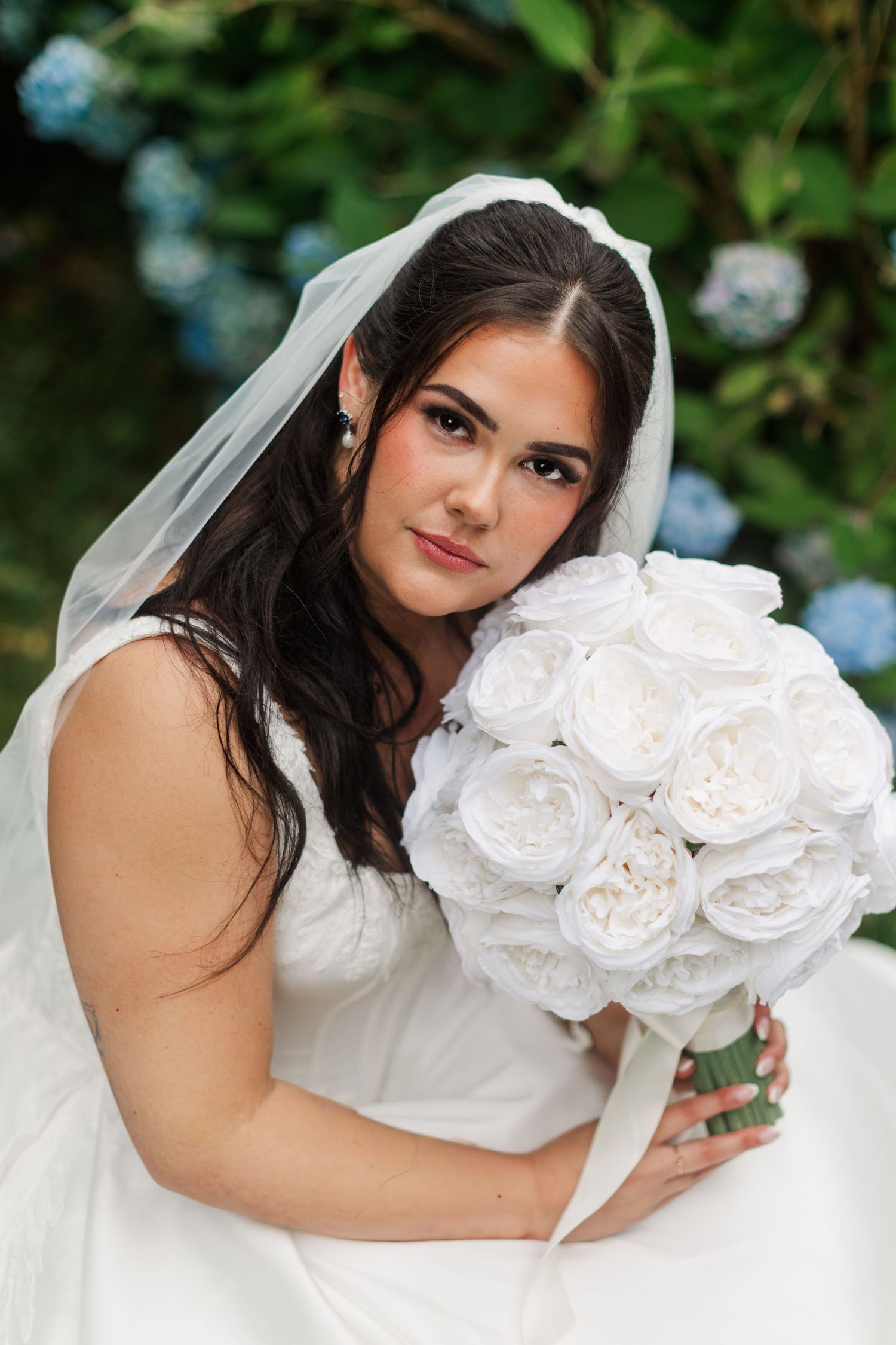 Bride in a white wedding gown and sheer veil holds a bouquet of white peonies while sitting in front of blue hydrangeas.