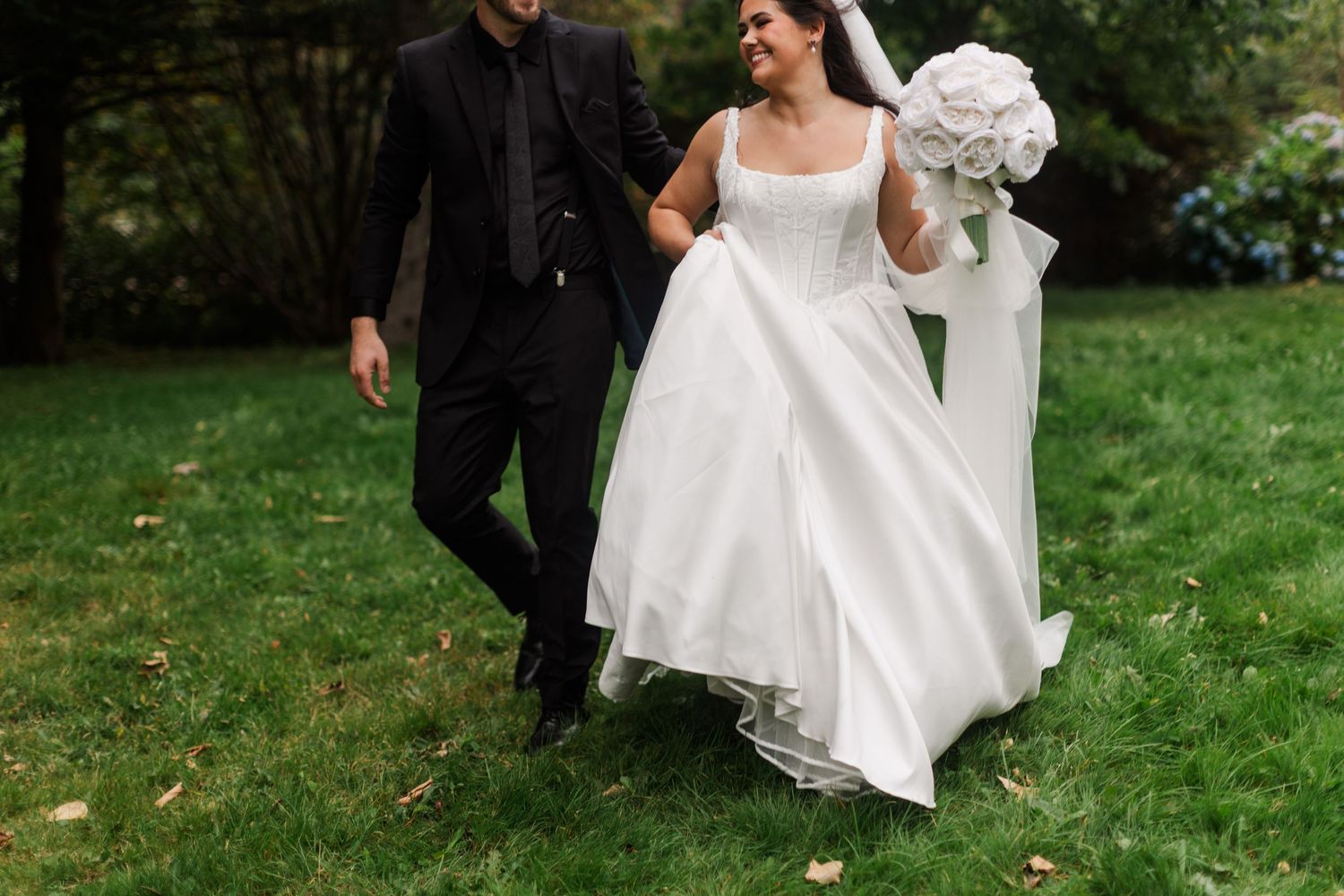 Bride in a flowing white gown holding a white bouquet walks hand in hand with a groom in a black suit across a green lawn.