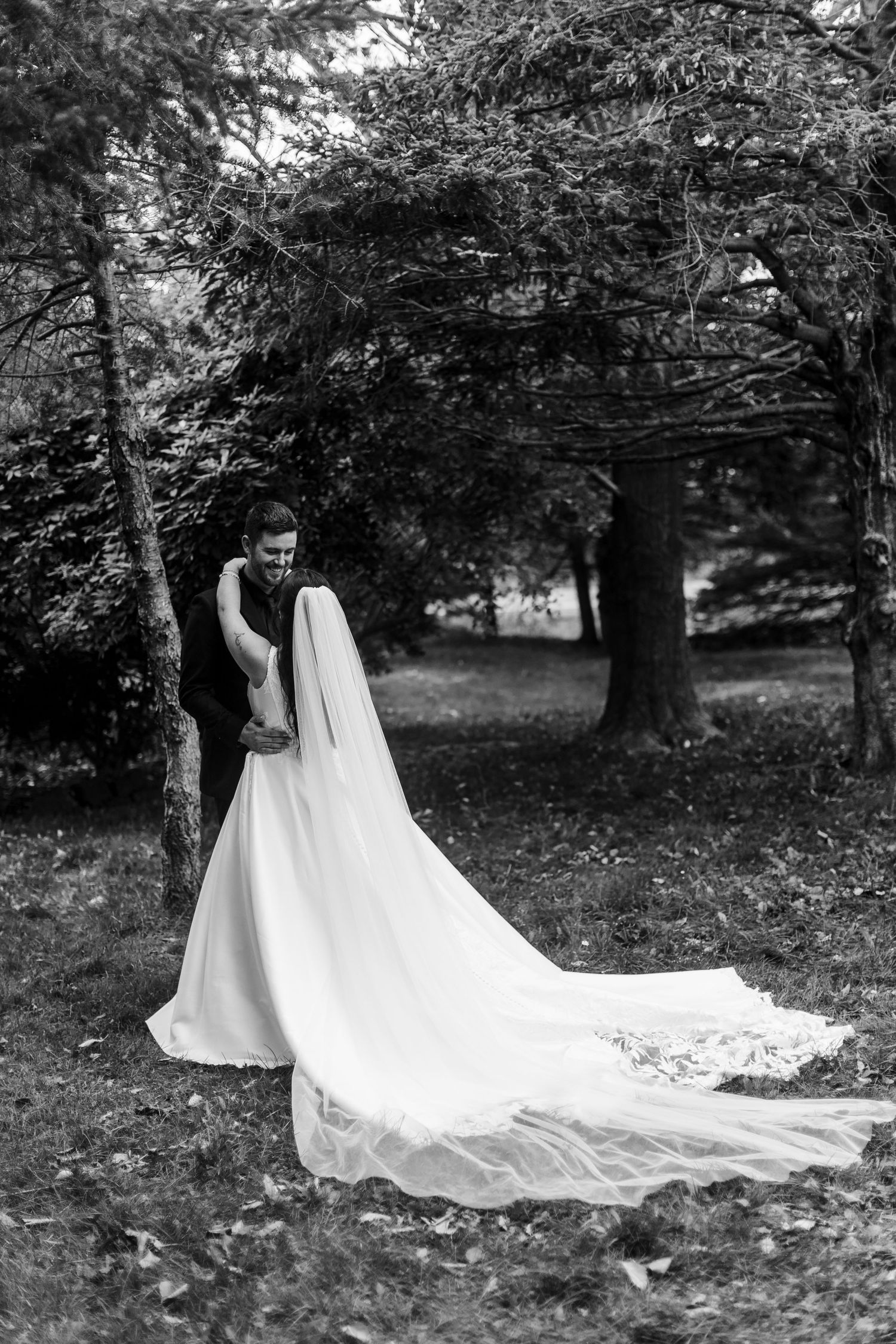 Black and white photo of a bride in a long gown and veil embracing her groom beneath tall trees in a peaceful park.
